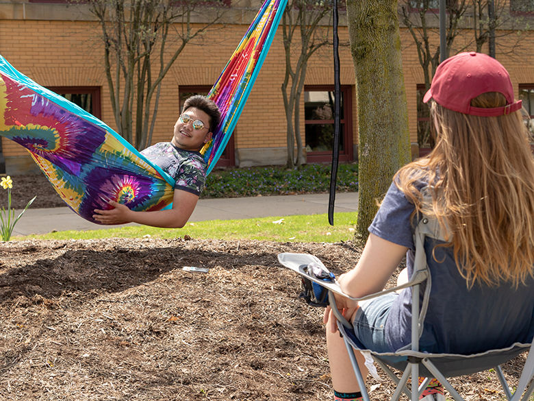 Student in a hammock talking to a student seated in folding chair